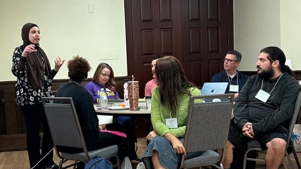A table of attendees listen to a standing speaker.